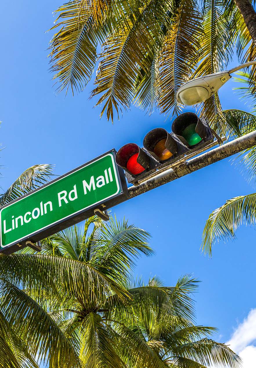 Señal de tránsito indicando "Lincoln Rd Mall" con semáforo al lado, rodeada de palmeras y cielo azul claro