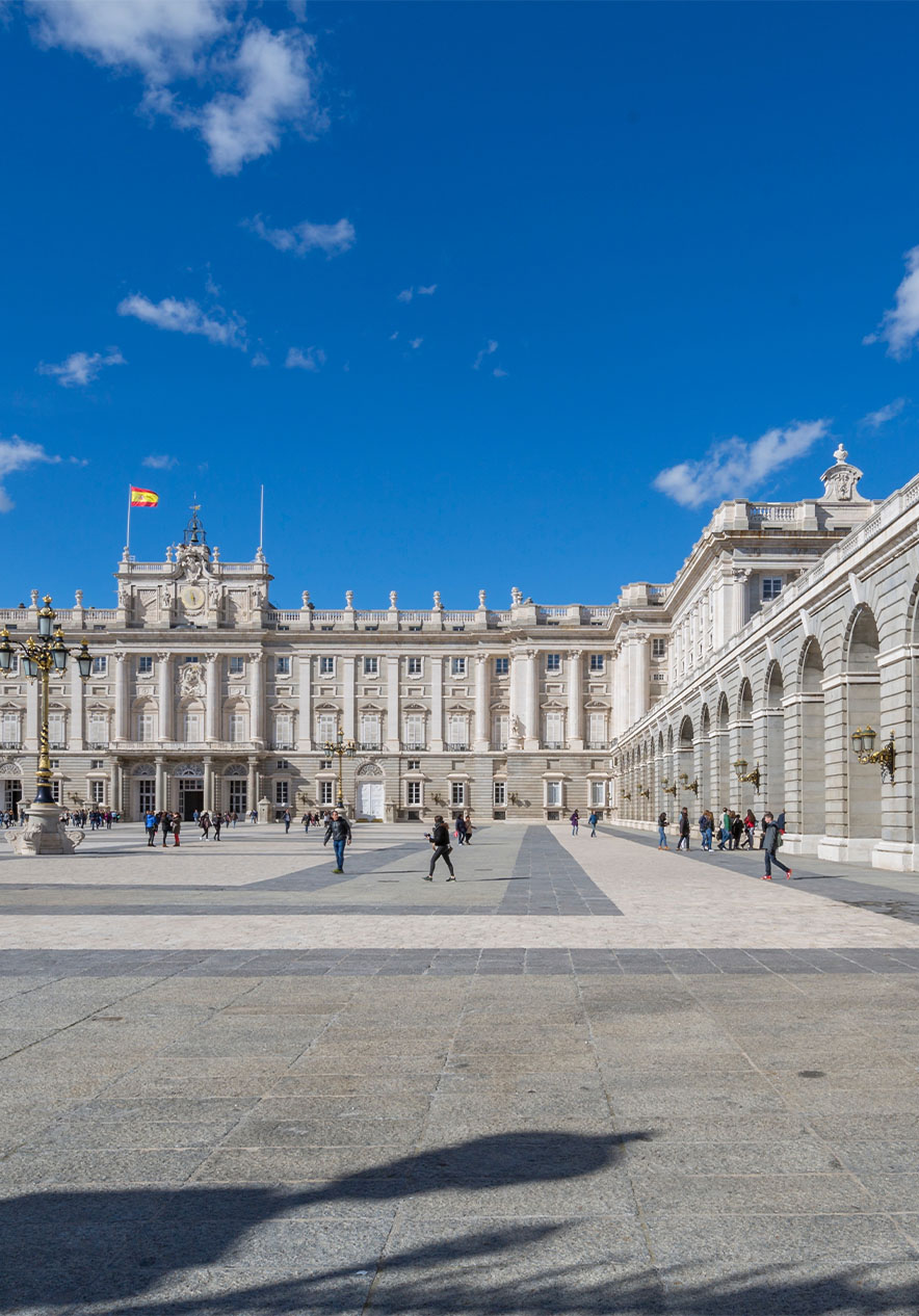 Patio del Palacio Real, con edificios de piedra formando un cuadrado, personas y la bandera de España