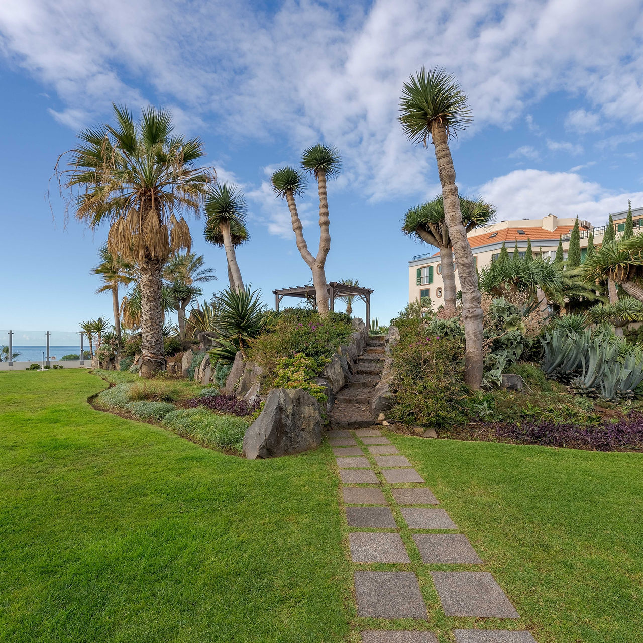 Paisaje de un jardín exuberante, con diversas especies de plantas, césped verde ordenado y frente al mar