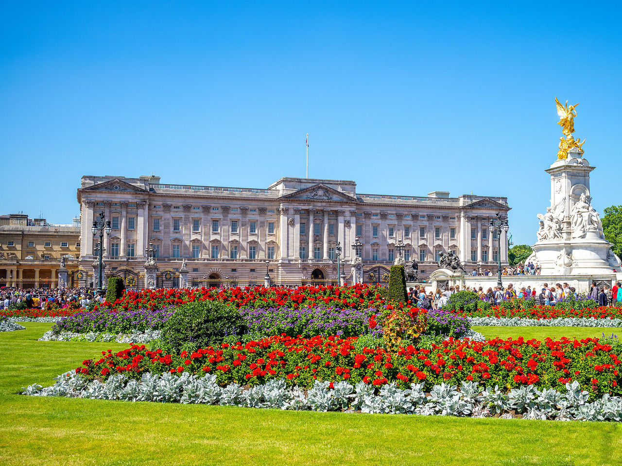 La imponente fachada del Palacio de Buckingham en Londres, con una estatua dorada y un jardín florecido en frente