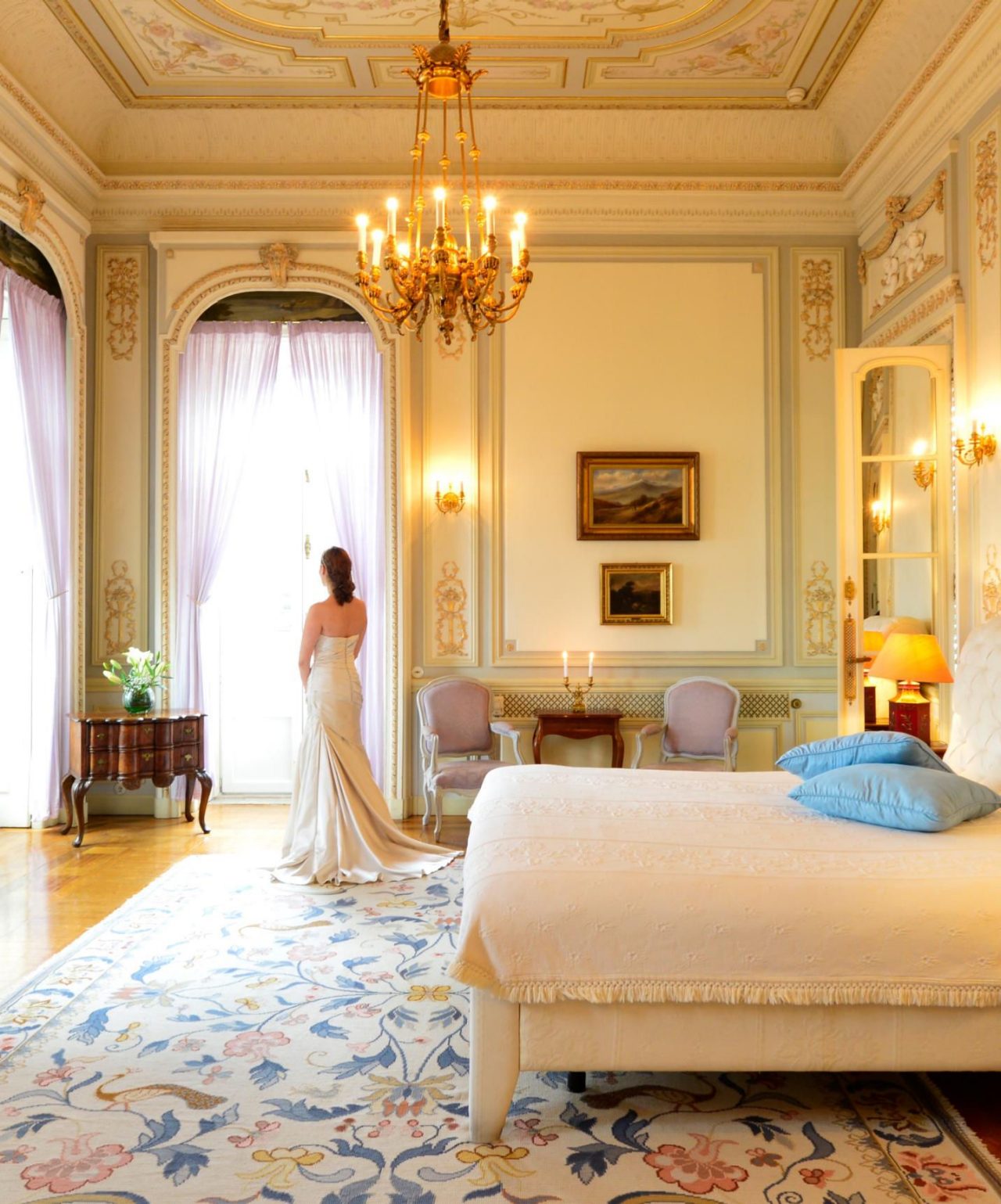 Novia se prepara para su boda en una elegante habitación de hotel, con paredes adornadas con detalles dorados y un candelabro de cristal