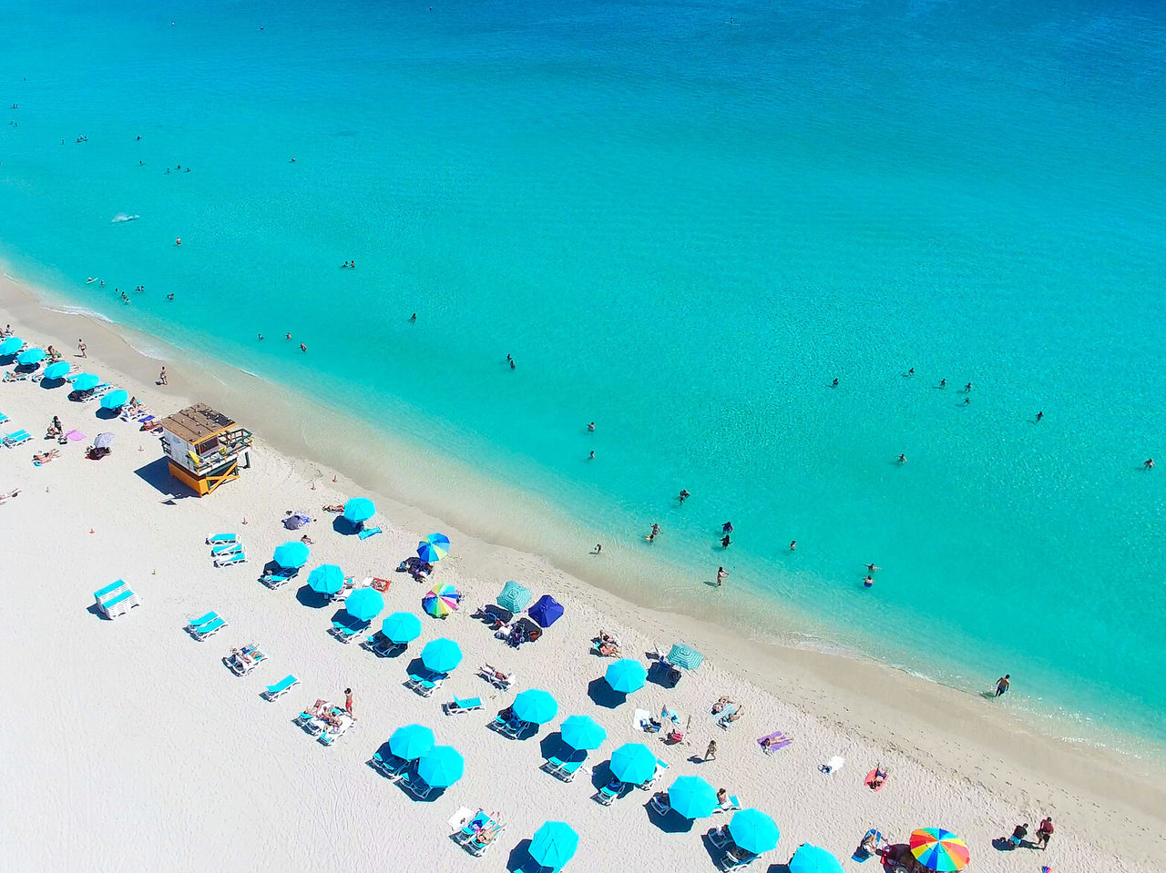 Praia de Miami Beach, Florida, com areia branca, água do mar azul turquesa e, pessoas a nadar e a apanhar sol nas cadeiras
