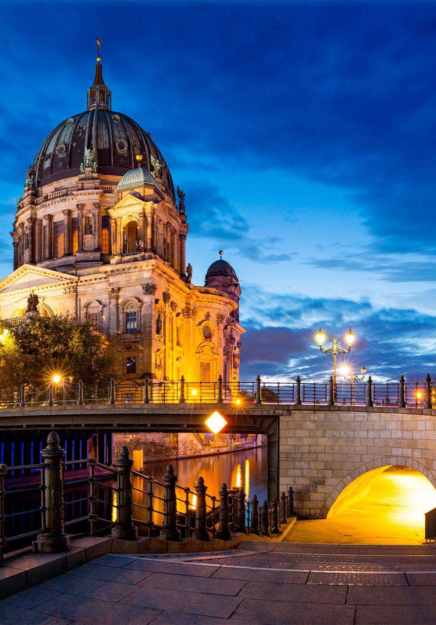 Catedral de Berlim iluminada ao anoitecer, com uma ponte e edifícios modernos ao lado, refletidos no rio abaixo