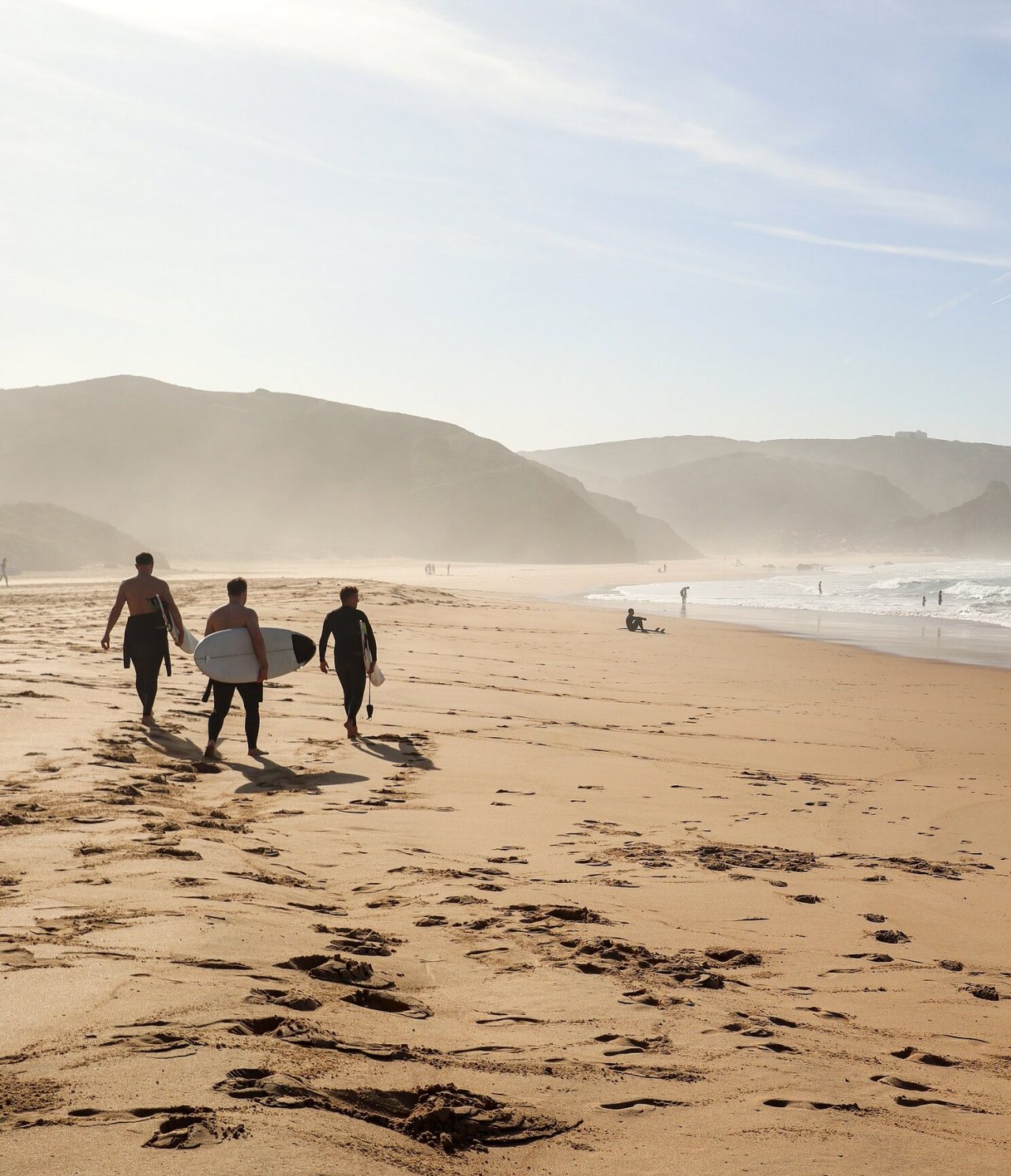 Surfistas a carregar a sua prancha de surf, numa praia na costa do Algarve, com ondas e rochas no fundo
