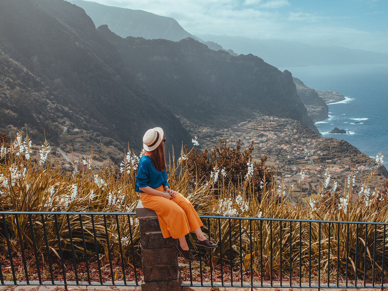 Mulher sentada num muro aprecia a vista para uma fajã na Ilha da Madeira, com o mar a contorná-la