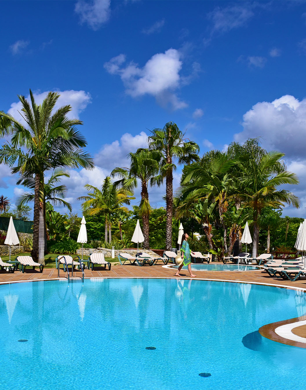 Outdoor pools with sun loungers, umbrellas, and palm trees, with a lady walking and the building on the side