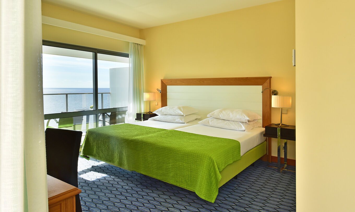 The Suite Sea View of Pestana Promenade has a double bed with a green bedspread and a large window to the balcony