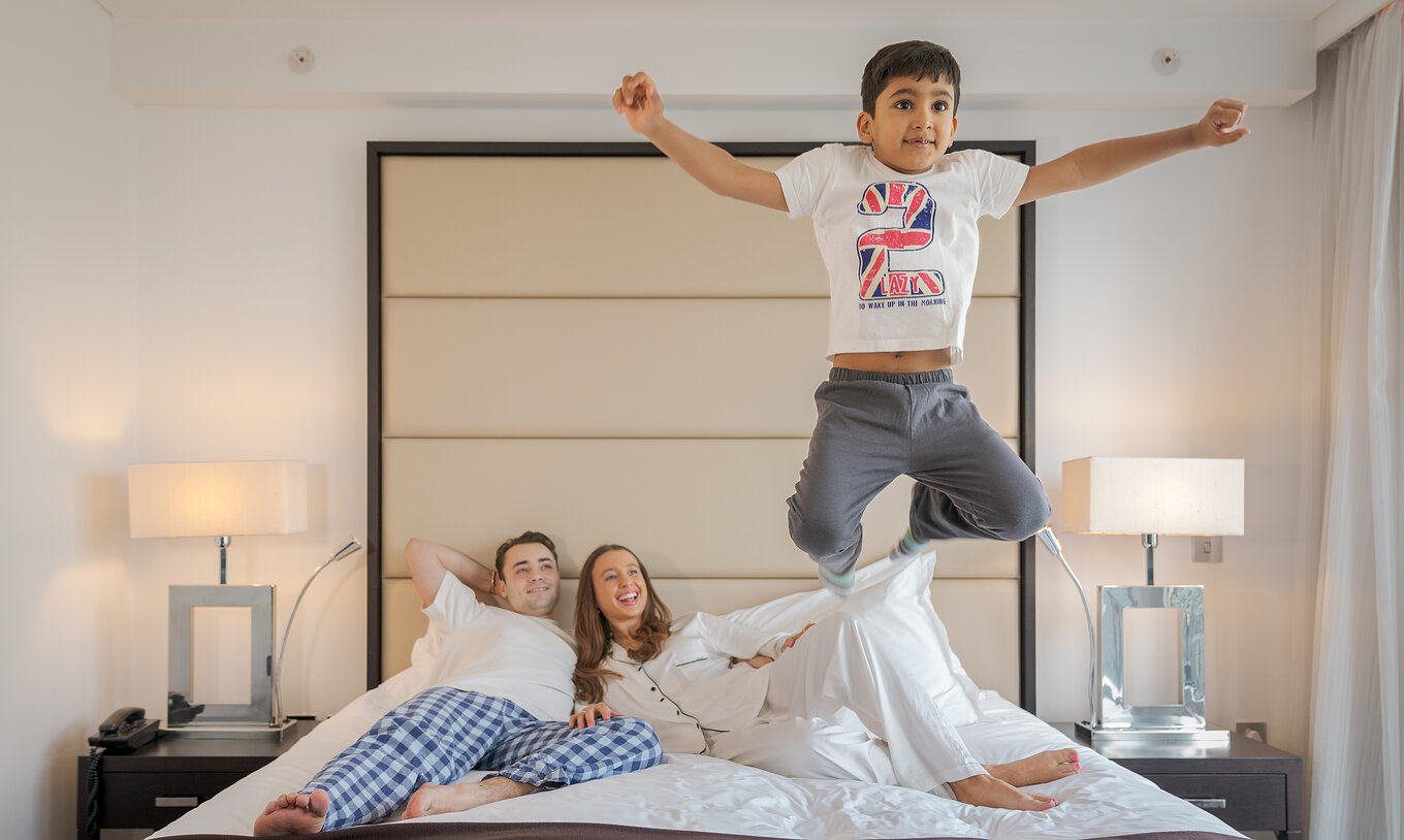 The Deluxe Family at Pestana Chelsea Bridge has a double bed with a family lying down and a child jumping