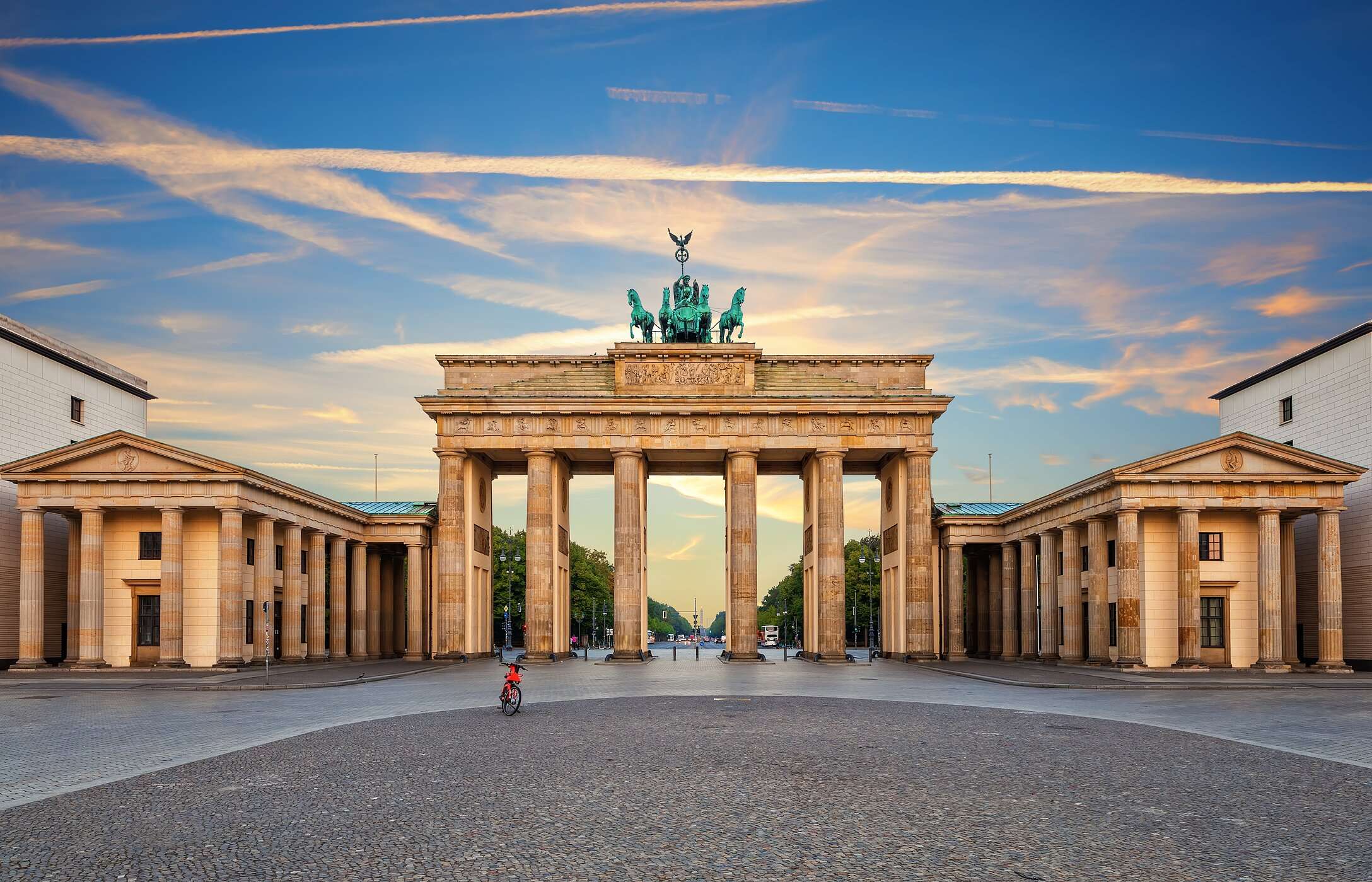 Paisaje urbano de Berlín, con la icónica Puerta de Brandeburgo, bajo un cielo azul y la avenida Unter den Linden al fondo.