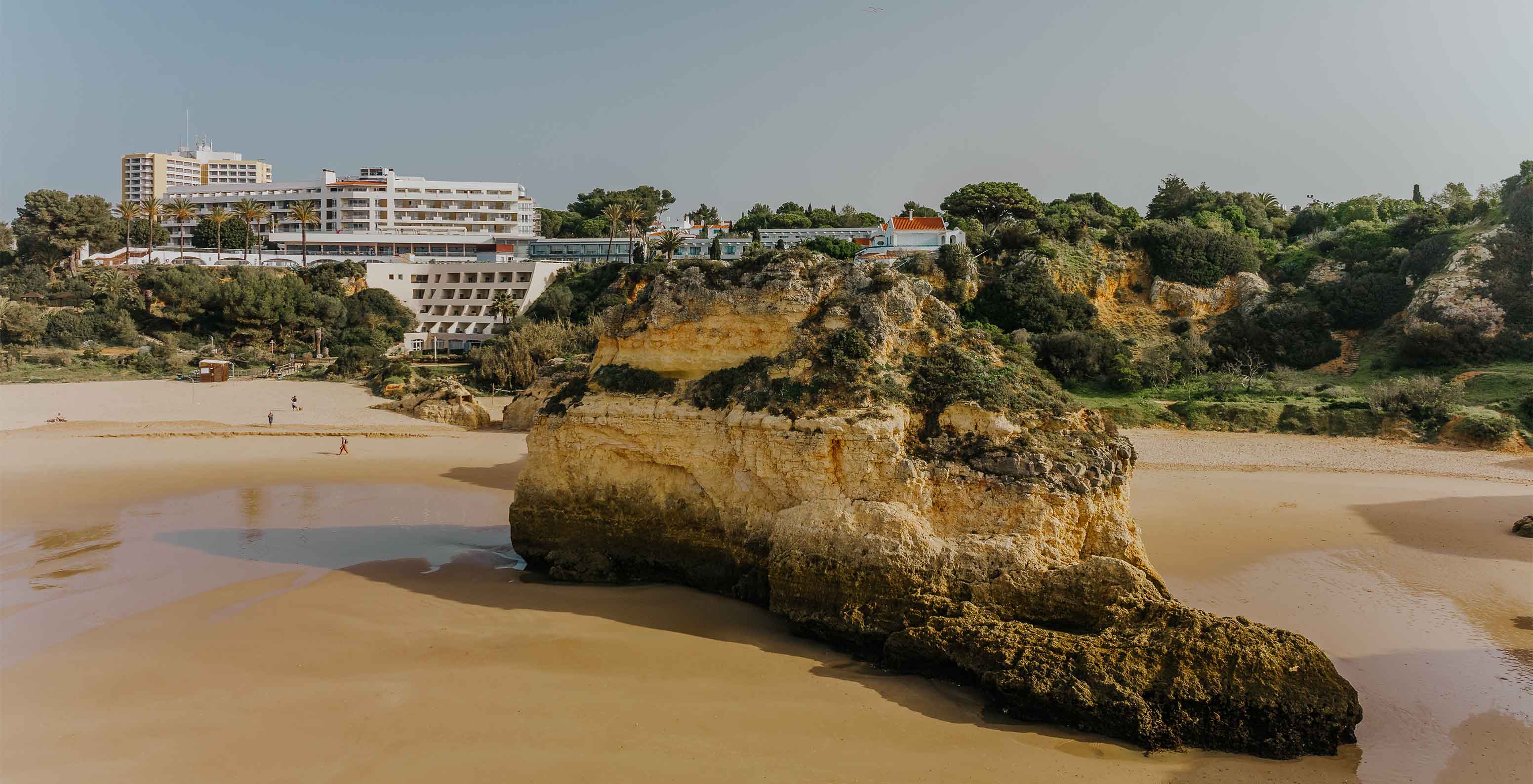 Playa de Alvor con rocas, arena, vegetación, el acantilado y edificios al fondo, con cielo despejado
