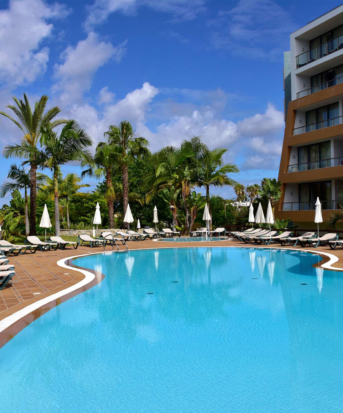 Buitenzwembaden met ligbedden, parasols en palmbomen, met het hotelgebouw aan de zijkant