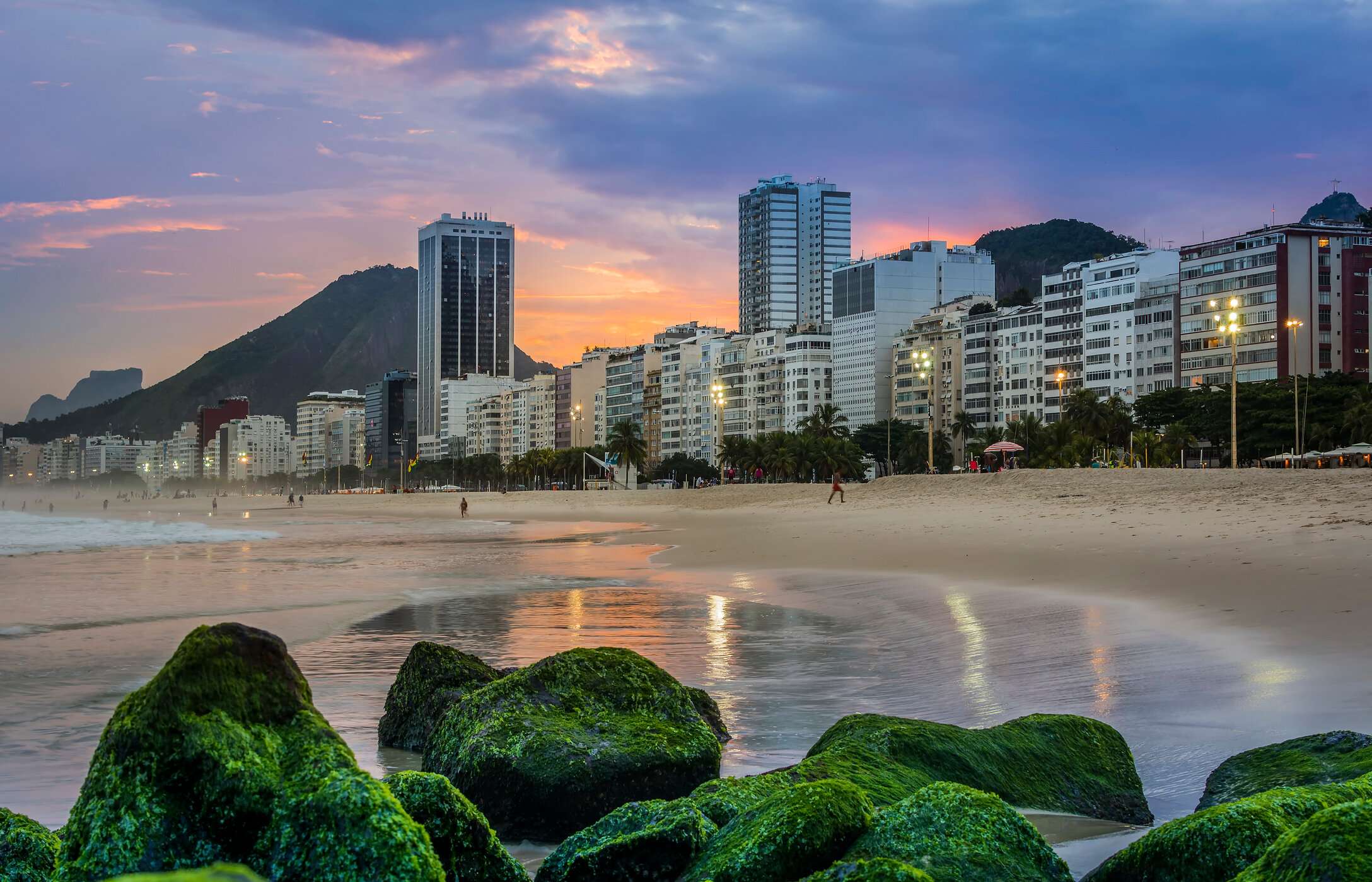Praia de Copacabana, com areias douradas, ondas do mar e um calçadão movimentado, cercado por prédios e montanhas ao fundo