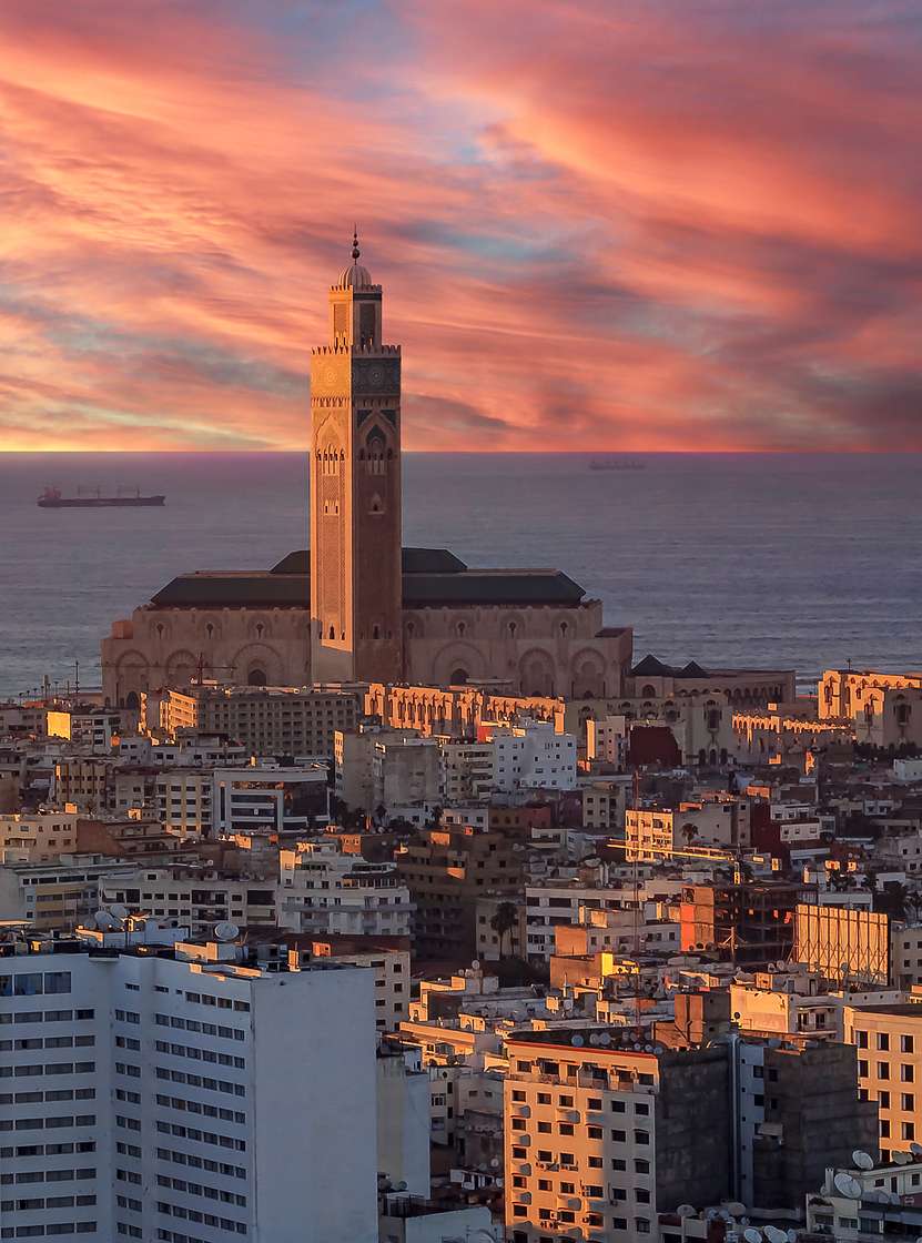Vista aérea sobre Casablanca, com a luz do pôr-do-sol a iluminar os vários prédios, o céu no horizonte sobre o mar e a torre