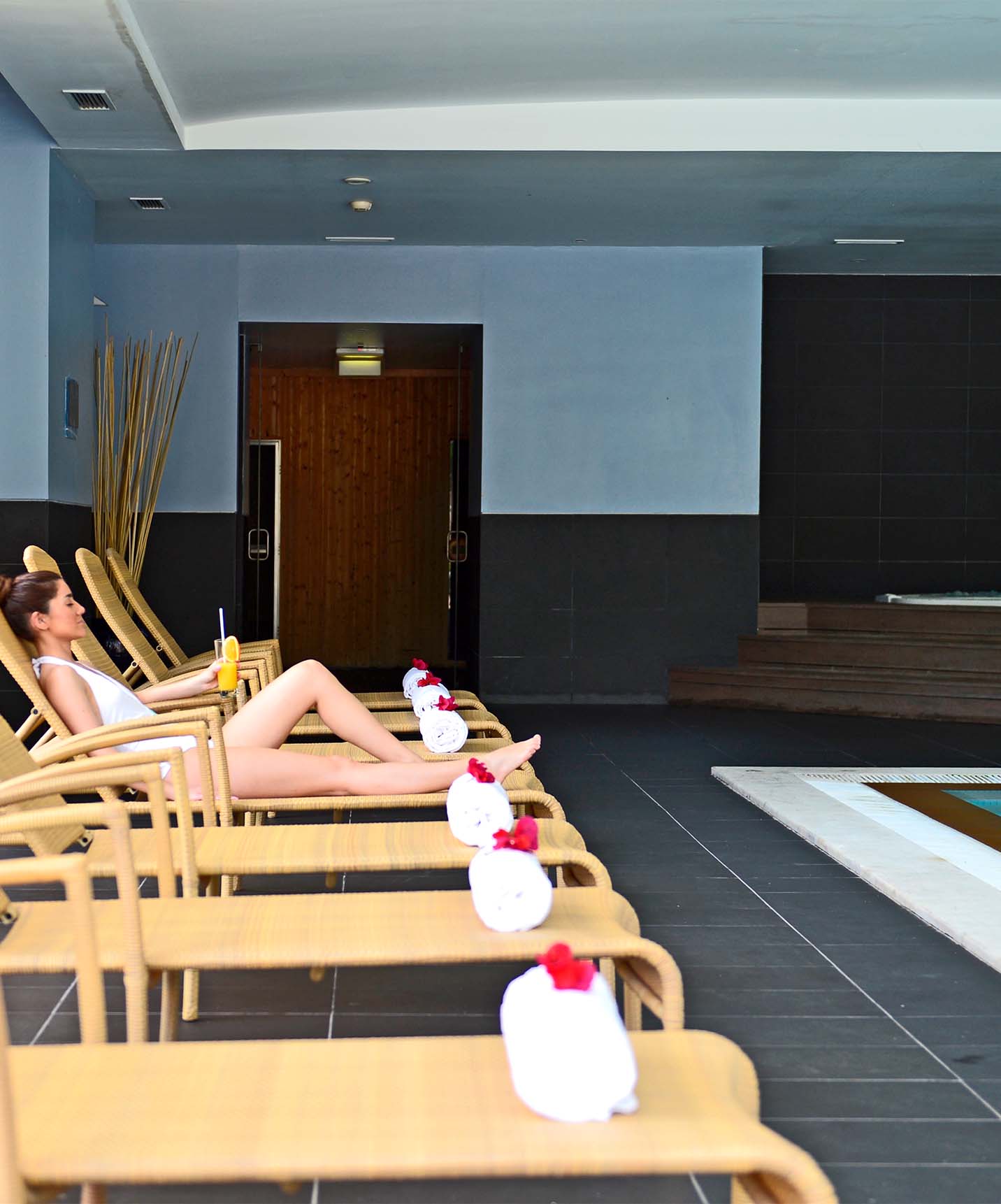 Piscina interior com escadas metálicas e jacuzzi, com espreguiçadeiras com toalhas e uma senhora deitada