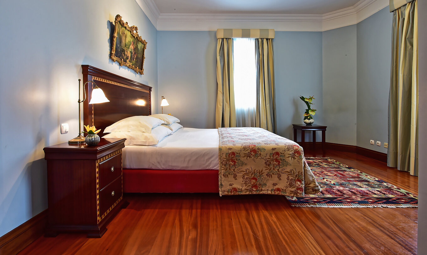A Suite Miramar do Pestana Miramar tem uma cama de casal com uma colcha às flores e uma mesa de cabeceira em madeira escura