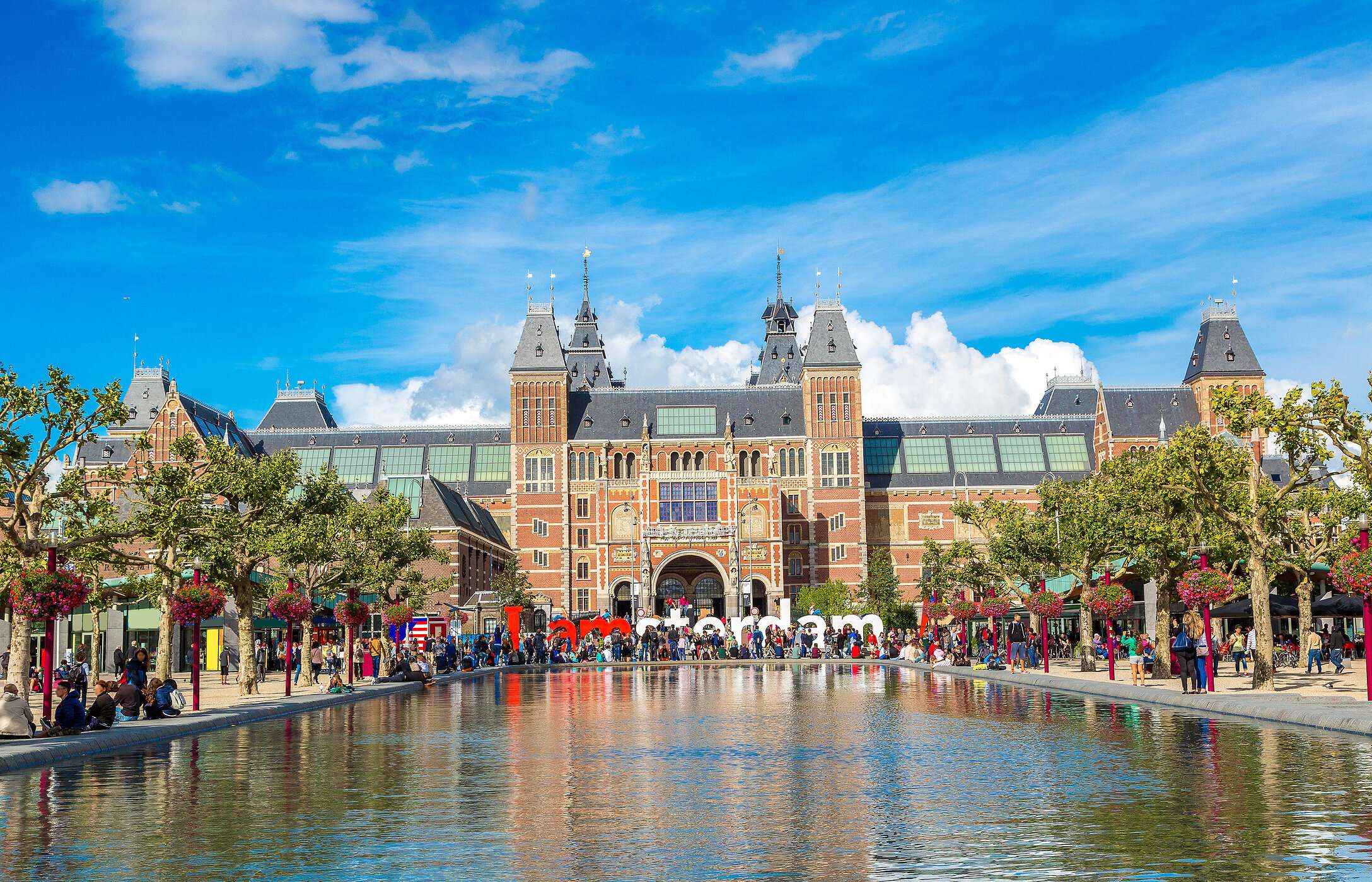 Fachada do famoso Rijksmuseum em Amsterdão, na Holanda, com várias pessoas a passear, árvores e um lago em primeiro plano