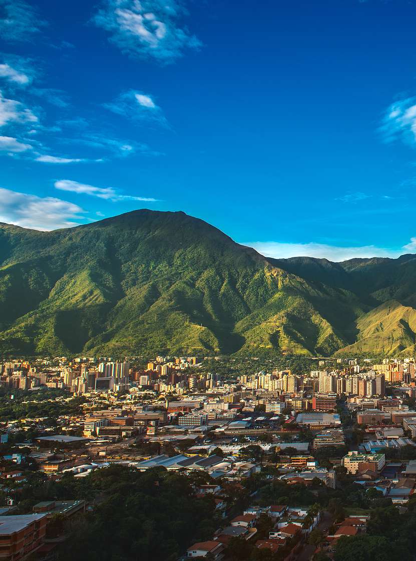 Vista aérea da cidade de Caracas, com prédios altos, contrastando com a natureza montanhosa, e o céu azul