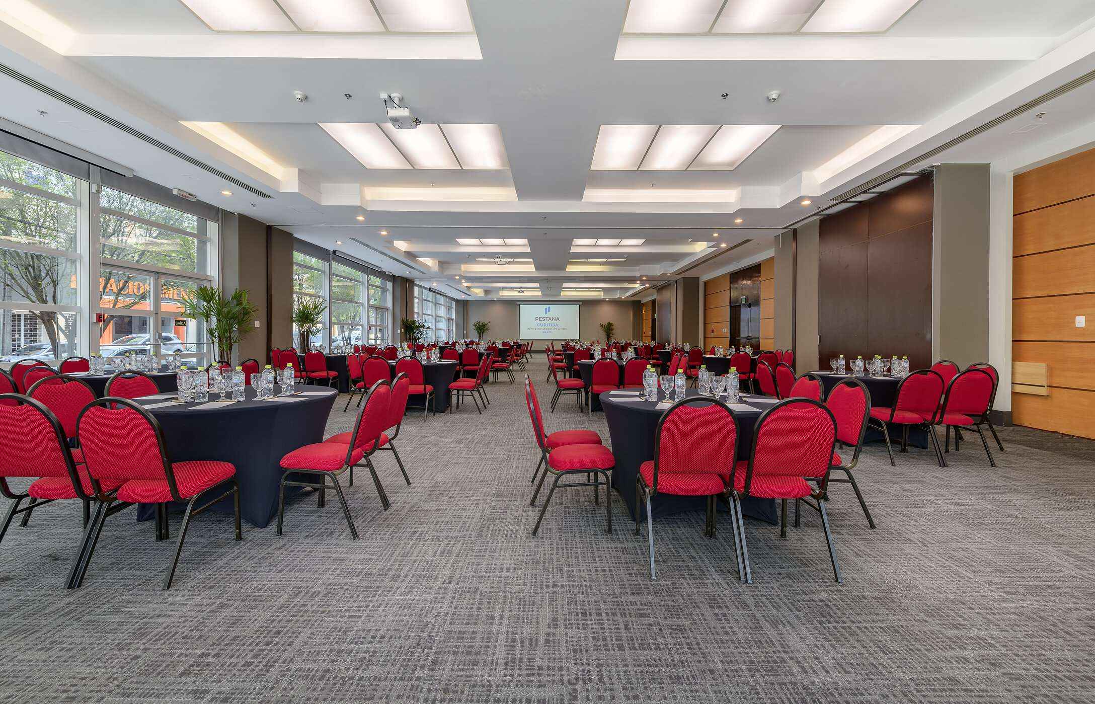 Sala para reuniões ou conferências, com várias mesas redondas com cadeiras vermelhas, copos e garrafas de água