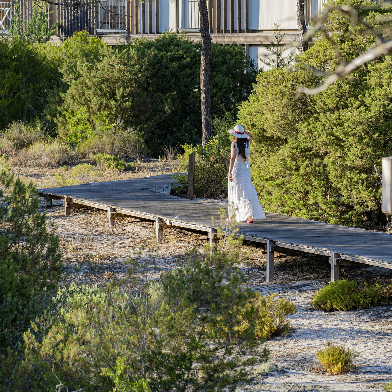 Wooden walkway at Pestana Tróia Eco Resort, where a woman in a white dress and hat strolls between the villas
