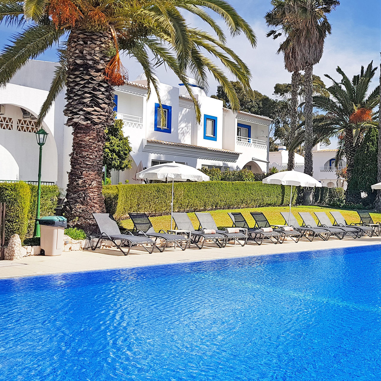 Outdoor pool at a hotel in Carvoeiro, with loungers, sunshades, palm trees, and villas behind