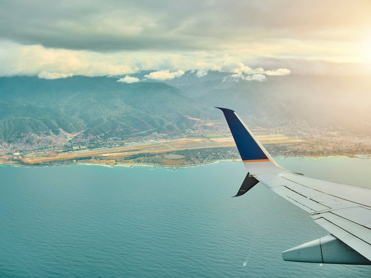 Aerial view of a plane wing flying over an ocean, with a city in the background, surrounded by mountains